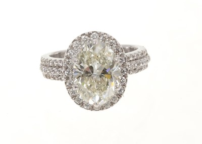 Lot 408 - Diamond halo cluster ring with an oval mixed-cut diamond weighing approximately 3cts, surrounded by a border of round brilliant cut diamonds with diamond set shoulders on a white gold shank.