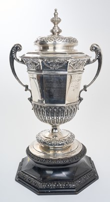 Lot 276 - Large, Edwardian silver twin handled trophy cup of octagonal form, with fluted and floral decoration, engraved presentation inscription and separate domed cover, on a carved ebonised plinth (L...