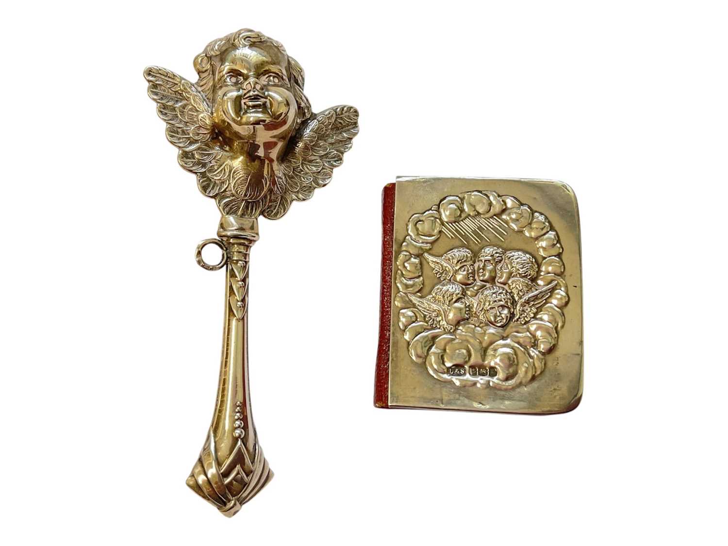 Lot 134 - Silver baby rattle with cupid head and wings decoration, 12cm, together with a silver mounted miniature prayer book decorated with Reynold's Angels (2)