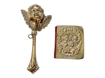 Lot 134 - Silver baby rattle with cupid head and wings decoration, 12cm, together with a silver mounted miniature prayer book decorated with Reynold's Angels (2)