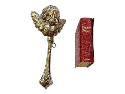 Lot 134 - Silver baby rattle with cupid head and wings decoration, 12cm, together with a silver mounted miniature prayer book decorated with Reynold's Angels (2)