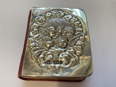 Lot 134 - Silver baby rattle with cupid head and wings decoration, 12cm, together with a silver mounted miniature prayer book decorated with Reynold's Angels (2)