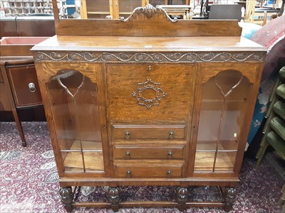 Lot 970 - 1930s oak bookcase with central fall flap and fitted interior and three drawers below flanked by two glazed doors on carved cup and cover supports